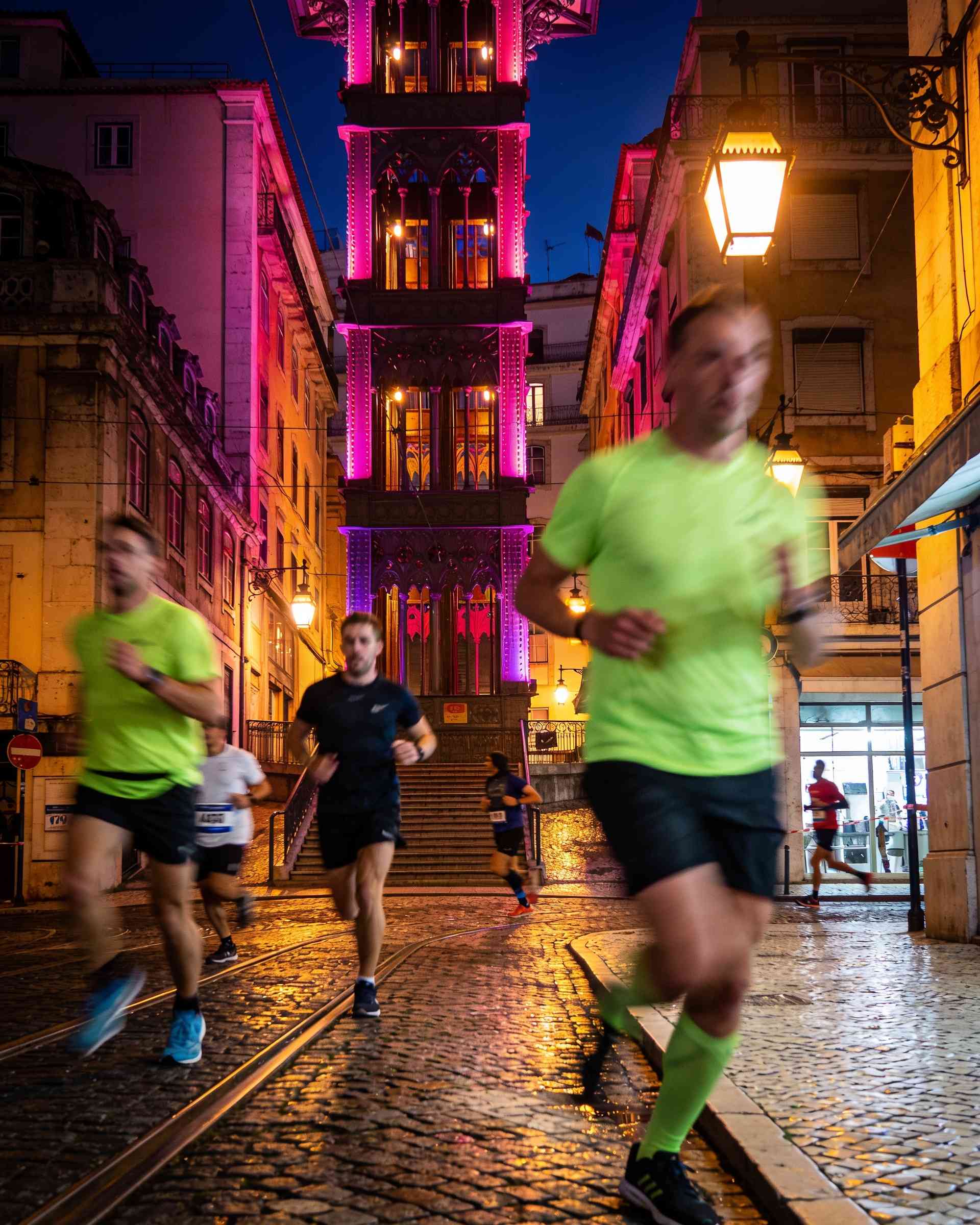 Santa Justa Lift in Lisbon at night, lit in magenta and violet, runners passing